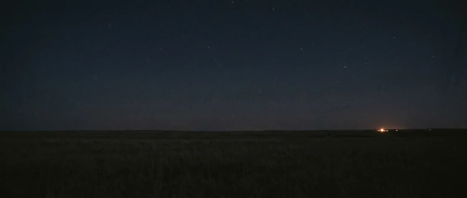 Night over open plains, a single distant town light low on the horizon under a dark sky.