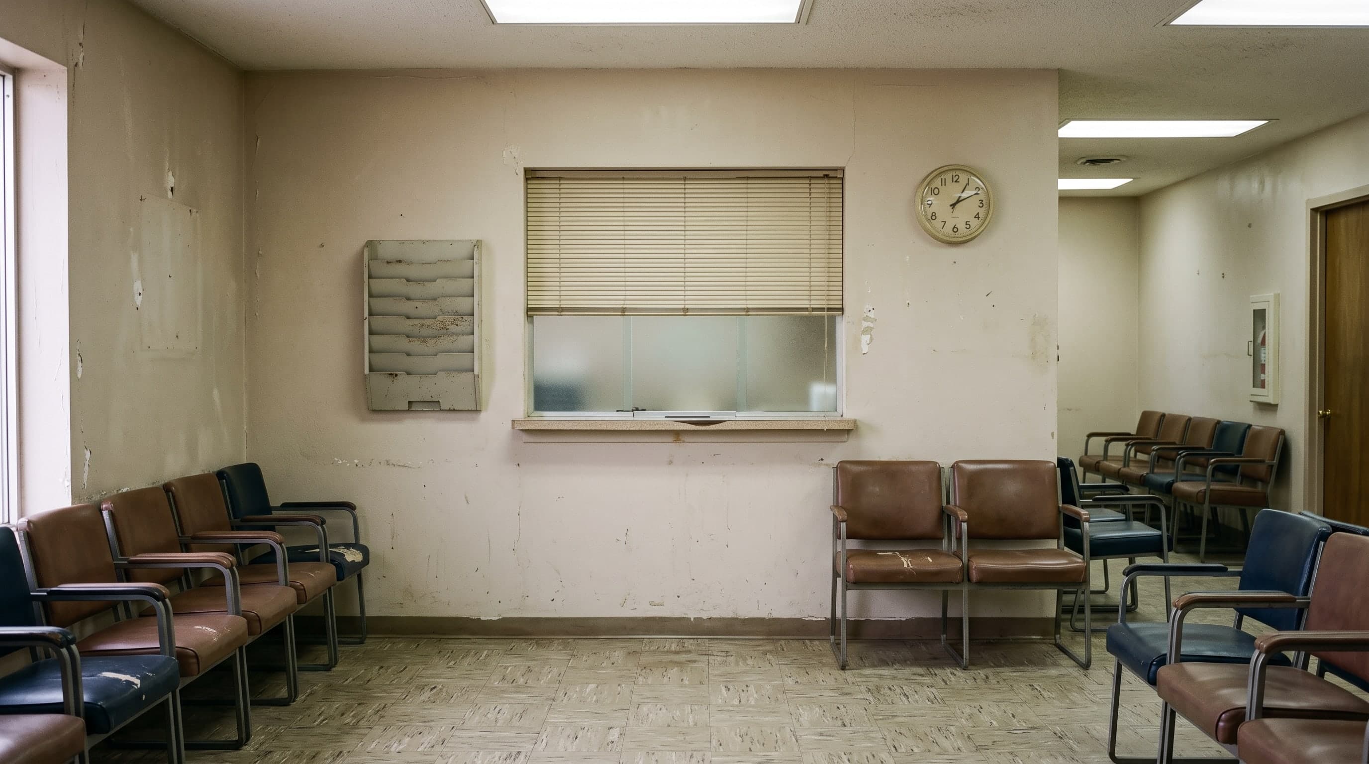 An empty waiting room with rows of worn vinyl chairs along the walls, a wall-mounted leaflet rack, a reception window with its blind drawn and a plain clock beside it, under fluorescent ceiling panels.