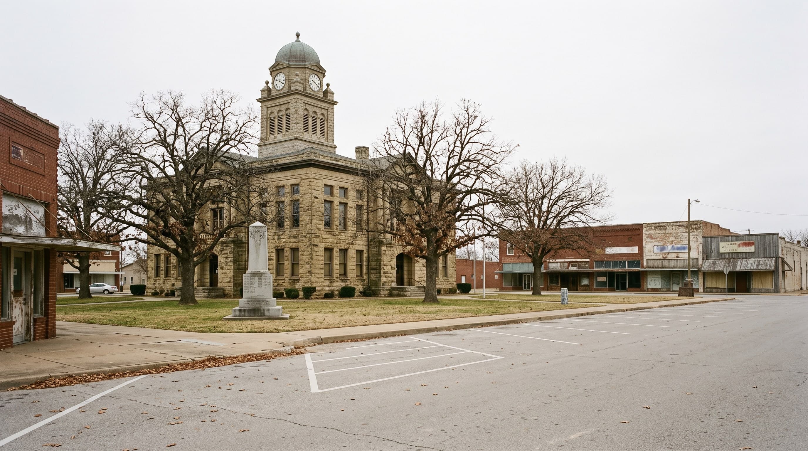 A limestone courthouse with a square clock tower on a small-town square, oaks and a memorial on the lawn.